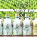 Row of olive oil bottles with olive tree branding arranged on a table against a green field backdrop