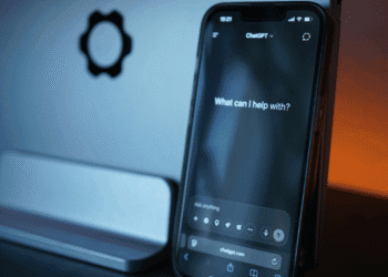 Smartphone displaying the ChatGPT interface with the prompt 'What can I help with?' in a blue-lit desk setup, gear icon visible on the wall behind it.