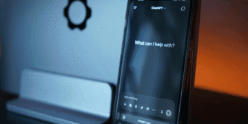 Smartphone displaying the ChatGPT interface with the prompt 'What can I help with?' in a blue-lit desk setup, gear icon visible on the wall behind it.