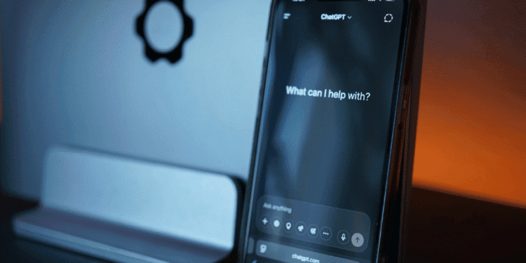 Smartphone displaying the ChatGPT interface with the prompt 'What can I help with?' in a blue-lit desk setup, gear icon visible on the wall behind it.
