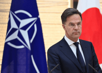 Man in a dark suit and tie speaks at a podium, with NATO flag on his left and the Japan flag on his right behind him.