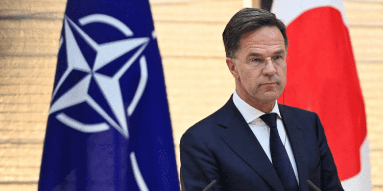 Delegação histórica da OTAN chega ao Japão para ampliar cooperação em defesa e segurança - ThingsOut Man in a dark suit and tie speaks at a podium, with NATO flag on his left and the Japan flag on his right behind him.