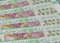 Row of colorful Japanese lottery tickets with green text and cartoon characters, stacked on a speckled surface. The tickets show a yellow mascot and playful illustrations.