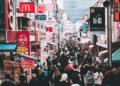 Crowded shopping street in Tokyo with colorful neon signs and storefronts on both sides.