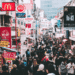 Crowded shopping street in Tokyo with colorful neon signs and storefronts on both sides.