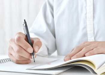 Person writes with a black ballpoint pen in a spiral notebook on a white desk, the page turned to reveal lines and a small orange highlight mark.