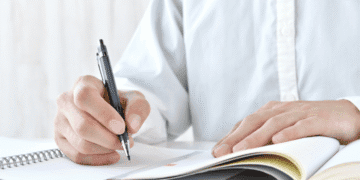 Person writes with a black ballpoint pen in a spiral notebook on a white desk, the page turned to reveal lines and a small orange highlight mark.