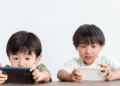 Two young boys sit at a table, each focused on a handheld device (smartphones) in their hands.