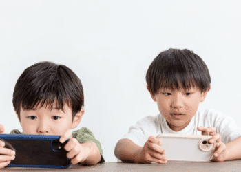 Two young boys sit at a table, each focused on a handheld device (smartphones) in their hands.