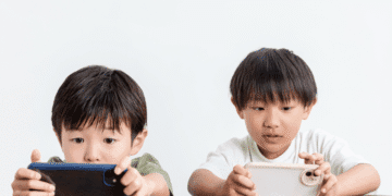 Two young boys sit at a table, each focused on a handheld device (smartphones) in their hands.