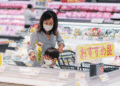 Mother and child wearing masks shop together in a bright supermarket, with a cart and shelves of produce in the background.