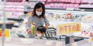 Mother and child wearing masks shop together in a bright supermarket, with a cart and shelves of produce in the background.