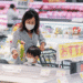 Mother and child wearing masks shop together in a bright supermarket, with a cart and shelves of produce in the background.
