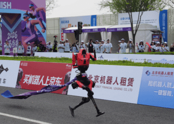 A red humanoid robot crosses a finish line on a race track as spectators watch from behind advertising boards and banners.