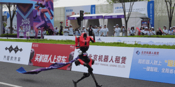 A red humanoid robot crosses a finish line on a race track as spectators watch from behind advertising boards and banners.