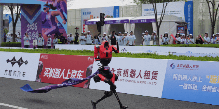 A red humanoid robot crosses a finish line on a race track as spectators watch from behind advertising boards and banners.