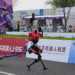 A red humanoid robot crosses a finish line on a race track as spectators watch from behind advertising boards and banners.