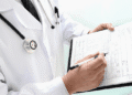 Doctor wearing a white coat and stethoscope writes on a medical form on a clipboard.