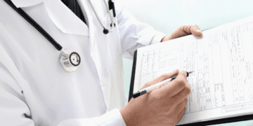 Doctor wearing a white coat and stethoscope writes on a medical form on a clipboard.