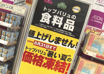 Supermarket display featuring a tall black-and-yellow sign announcing a price freeze for Topvalu food items, with shelves of canned goods on the left and boxed products on the right.