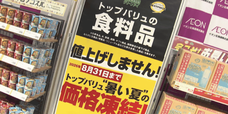 Supermarket display featuring a tall black-and-yellow sign announcing a price freeze for Topvalu food items, with shelves of canned goods on the left and boxed products on the right.