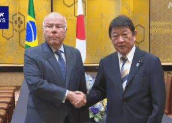 Two men in suits shake hands in a formal meeting room with Brazil and Japan flags behind them.
