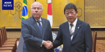 Two men in suits shake hands in a formal meeting room with Brazil and Japan flags behind them.