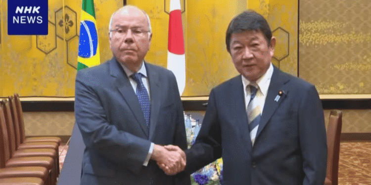 Two men in suits shake hands in a formal meeting room with Brazil and Japan flags behind them.