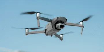 Drone in midair: a gray Mavic 2 quadcopter with forward-facing camera against a blue sky, propellers blurred from motion.