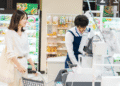 Governo japonês avalia reduzir imposto sobre alimentos para 1 por dois anos - ThingsOut Smiling woman with a shopping cart at the checkout while a cashier in a blue apron scans items at a grocery store register.