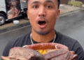 Influenciador americano é criticado após cuspir alimentos em vídeos durante viagem ao Japão - ThingsOut Young man with a shocked expression holding a massive smoked meat roast outdoors, with a food truck in the background.