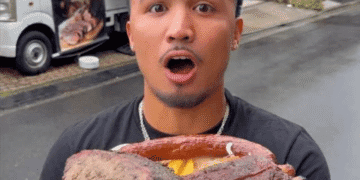 Young man with a shocked expression holding a massive smoked meat roast outdoors, with a food truck in the background.