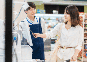 Female customer using a phone to pay at the checkout while a smiling cashier in a blue apron stands nearby.