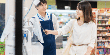 Female customer using a phone to pay at the checkout while a smiling cashier in a blue apron stands nearby.