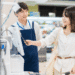 Female customer using a phone to pay at the checkout while a smiling cashier in a blue apron stands nearby.
