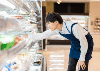 Man in a blue apron reaches into a grocery store refrigerated shelf to pick an item.