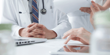 Medical team in a consultation, hands folded on a table with a laptop and patient forms.