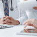 Medical team in a consultation, hands folded on a table with a laptop and patient forms.