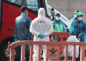 Medical responders in PPE stand beside a red emergency vehicle behind orange barriers, at a cordoned area.