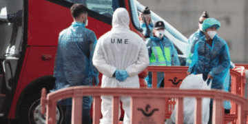 Medical responders in PPE stand beside a red emergency vehicle behind orange barriers, at a cordoned area.
