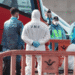 Medical responders in PPE stand beside a red emergency vehicle behind orange barriers, at a cordoned area.