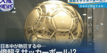 News broadcast screenshot showing a large gold metallic soccer ball with Japanese captions about a colossal ball