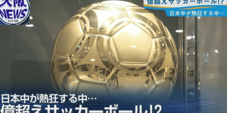 News broadcast screenshot showing a large gold metallic soccer ball with Japanese captions about a colossal ball