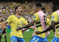 Brazil national team players celebrate a goal on the soccer field, wearing yellow jerseys and pink training bibs in front of a cheering crowd.