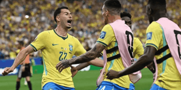 Brazil national team players celebrate a goal on the soccer field, wearing yellow jerseys and pink training bibs in front of a cheering crowd.