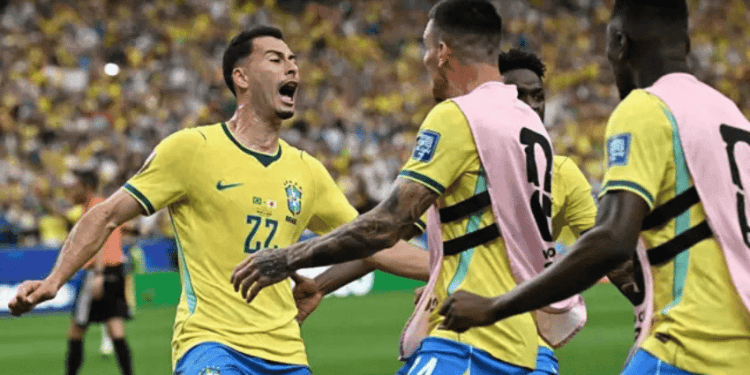 Brazil national team players celebrate a goal on the soccer field, wearing yellow jerseys and pink training bibs in front of a cheering crowd.