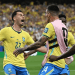 Brazil national team players celebrate a goal on the soccer field, wearing yellow jerseys and pink training bibs in front of a cheering crowd.
