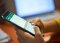 Golpe no WhatsApp usa SMS falso para assustar usuários e tentar roubar códigos de verificação - ThingsOut Person typing on a smartphone while a laptop sits blurred in the background.