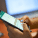 Golpe no WhatsApp usa SMS falso para assustar usuários e tentar roubar códigos de verificação - ThingsOut Person typing on a smartphone while a laptop sits blurred in the background.