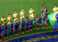 Brazil national team players and children stand in line with hands over their hearts on a soccer pitch, a flag bearer holding the Brazilian flag nearby
